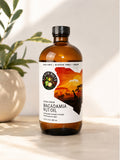 Elgon Nuts 16.9 fl oz macadamia nut oil bottle displayed on a sunlit countertop beside a ceramic vase with greenery.